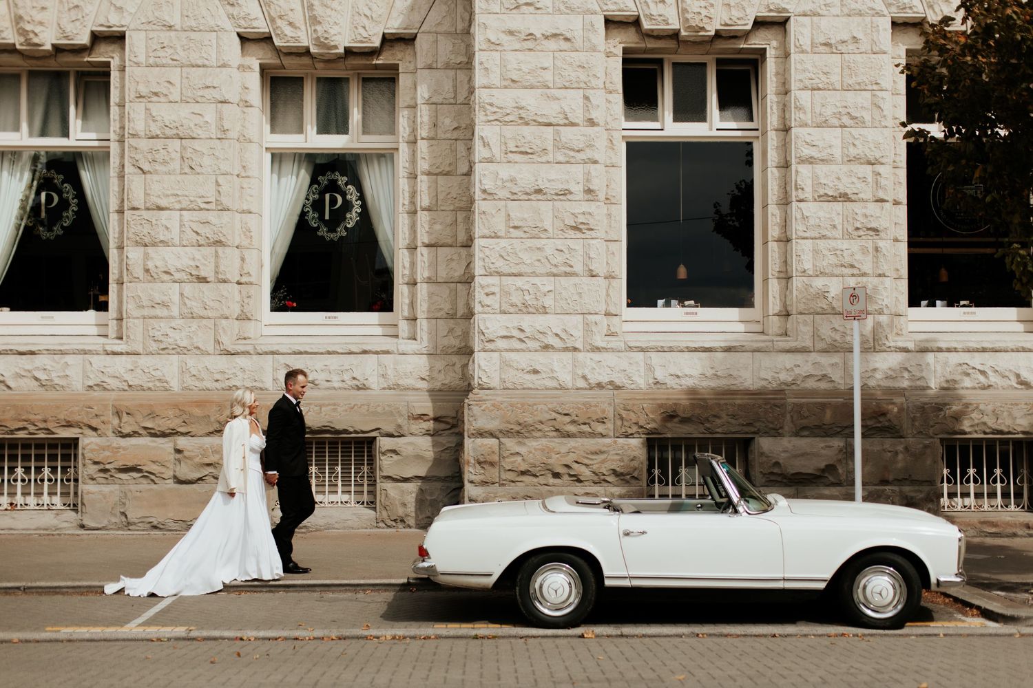 Classic white Mercedes convertible parked outside stone building with wedding couple in frame.