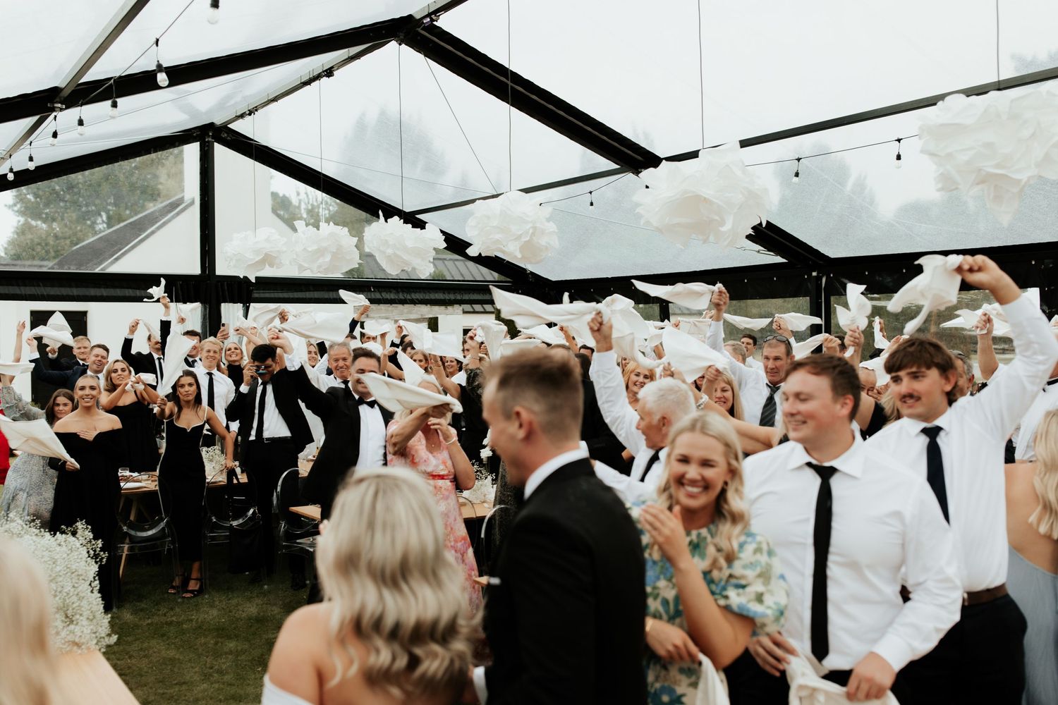 Wedding guests celebrate under a clear tent canopy during a lively reception party.