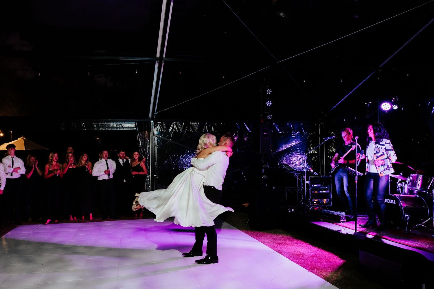 Romantic couple performing choreographed dance on illuminated purple dance floor with band in background.