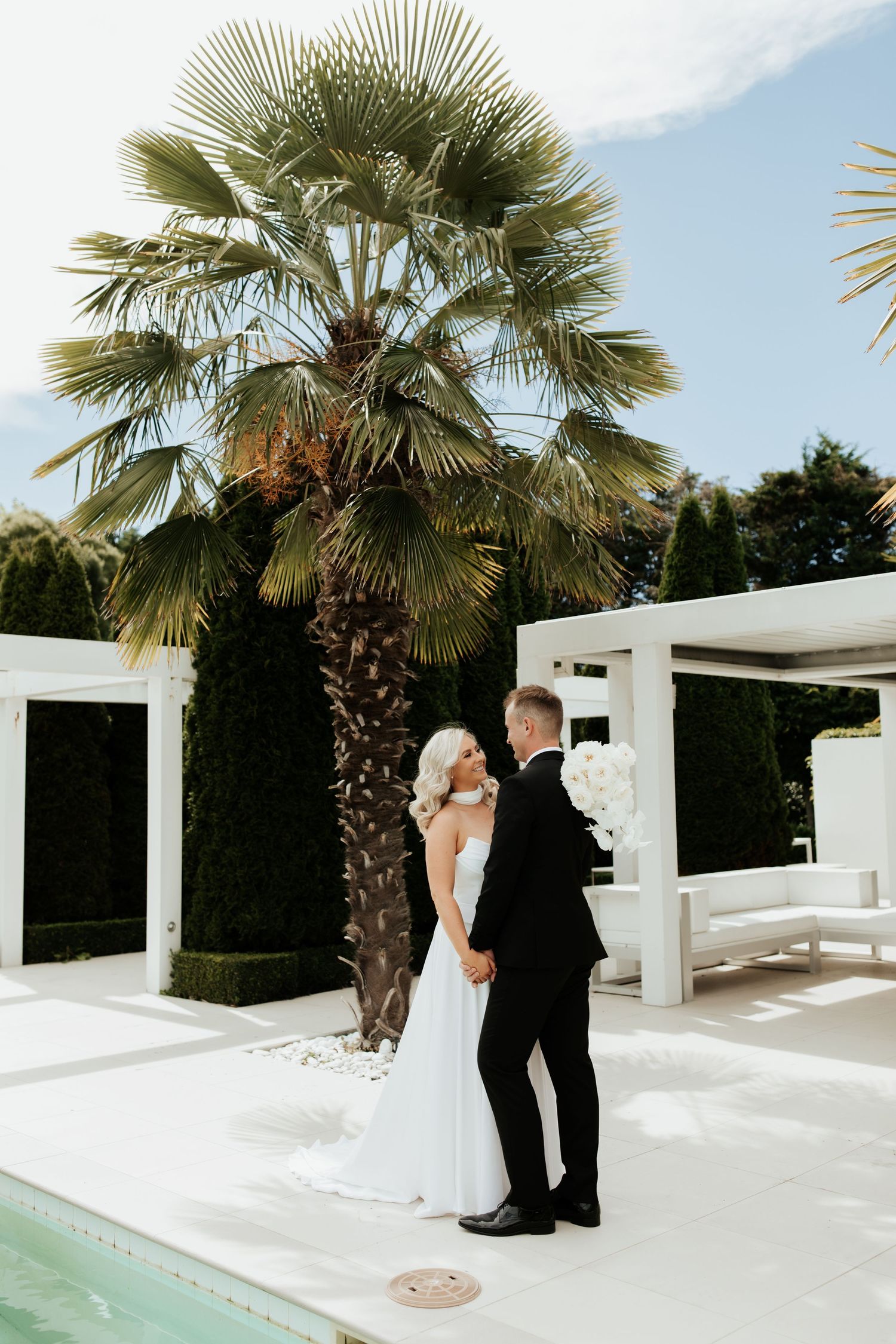 Elegant couple poses by palm tree and pool at modern white venue.
