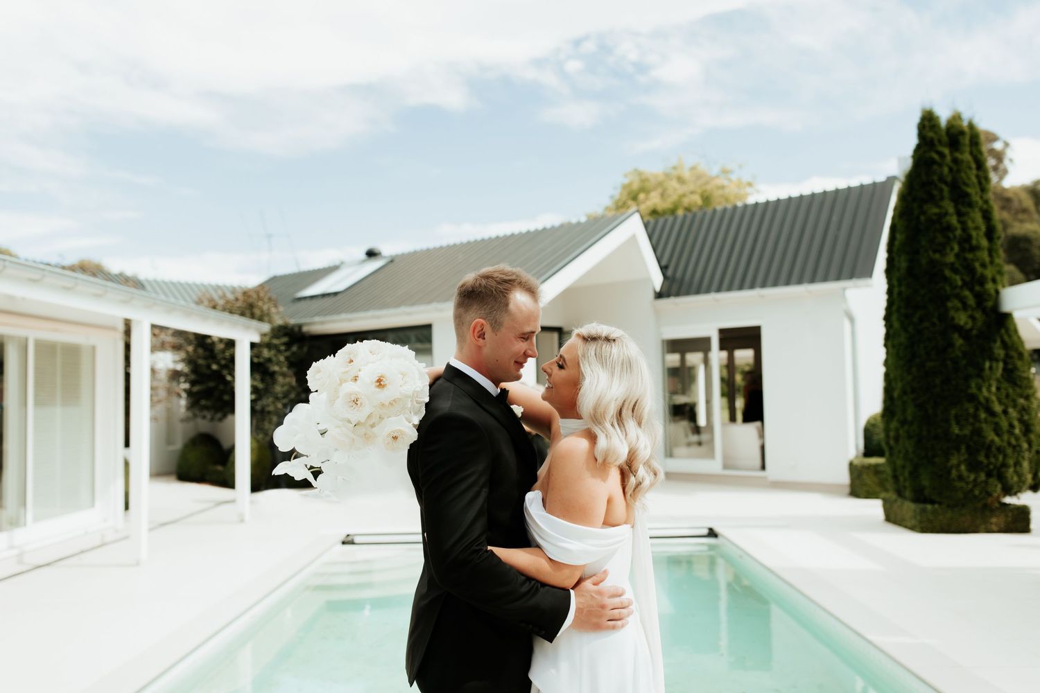 Newlyweds share intimate moment by pool with white buildings and orchid bouquet.