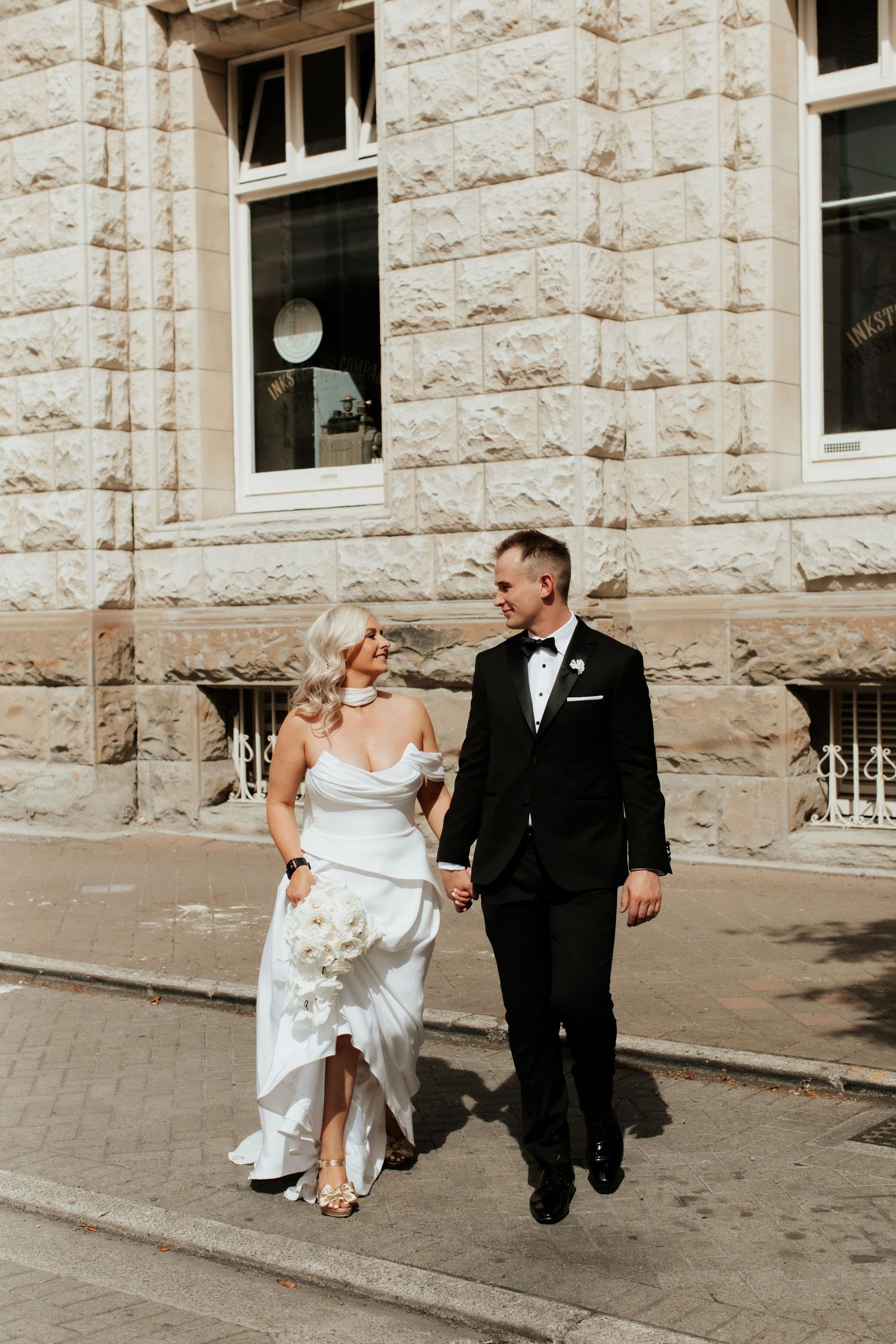 Elegantly dressed couple walking together outside a historic stone building on their wedding day.
