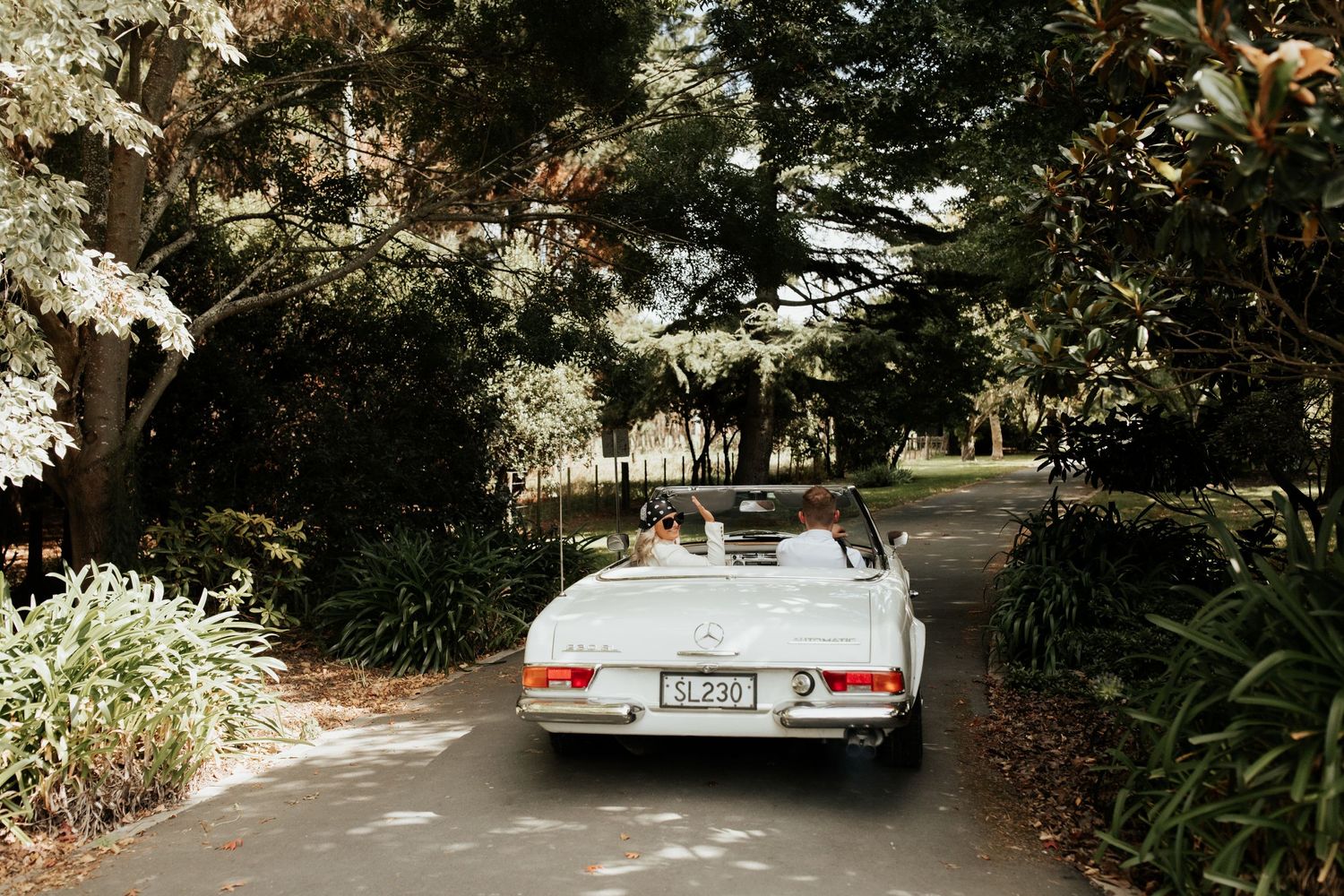 Vintage white Mercedes-Benz convertible driving through a lush garden setting.
