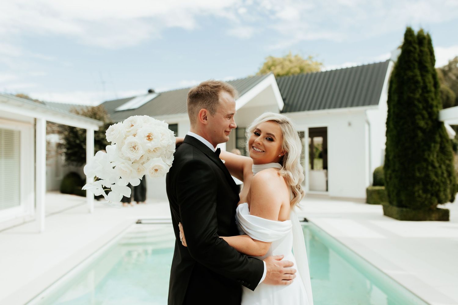 Couple in formal attire embracing by modern pool with white buildings and orchid bouquet.