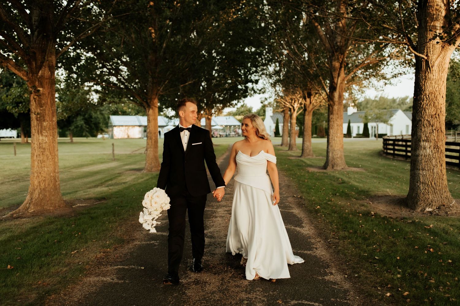 Newlyweds walk hand in hand through a tree-lined path during golden hour.
