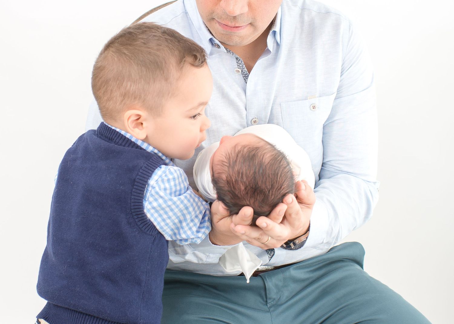 two year old brother about to kiss his little newborn baby brother on the face white dad is holding baby in an all white