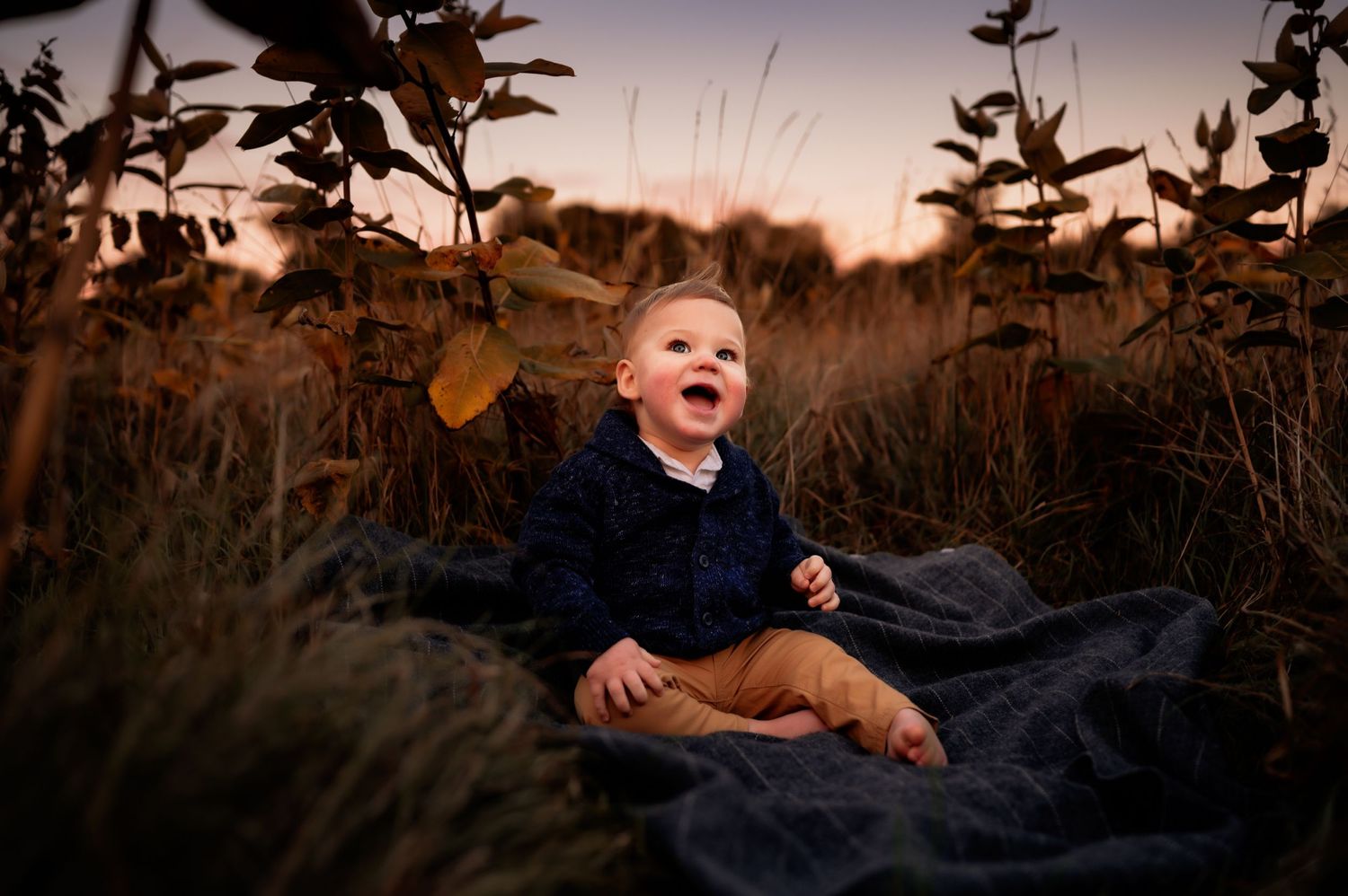 Joyful child sitting on a blanket in tall grass during sunset with golden light.