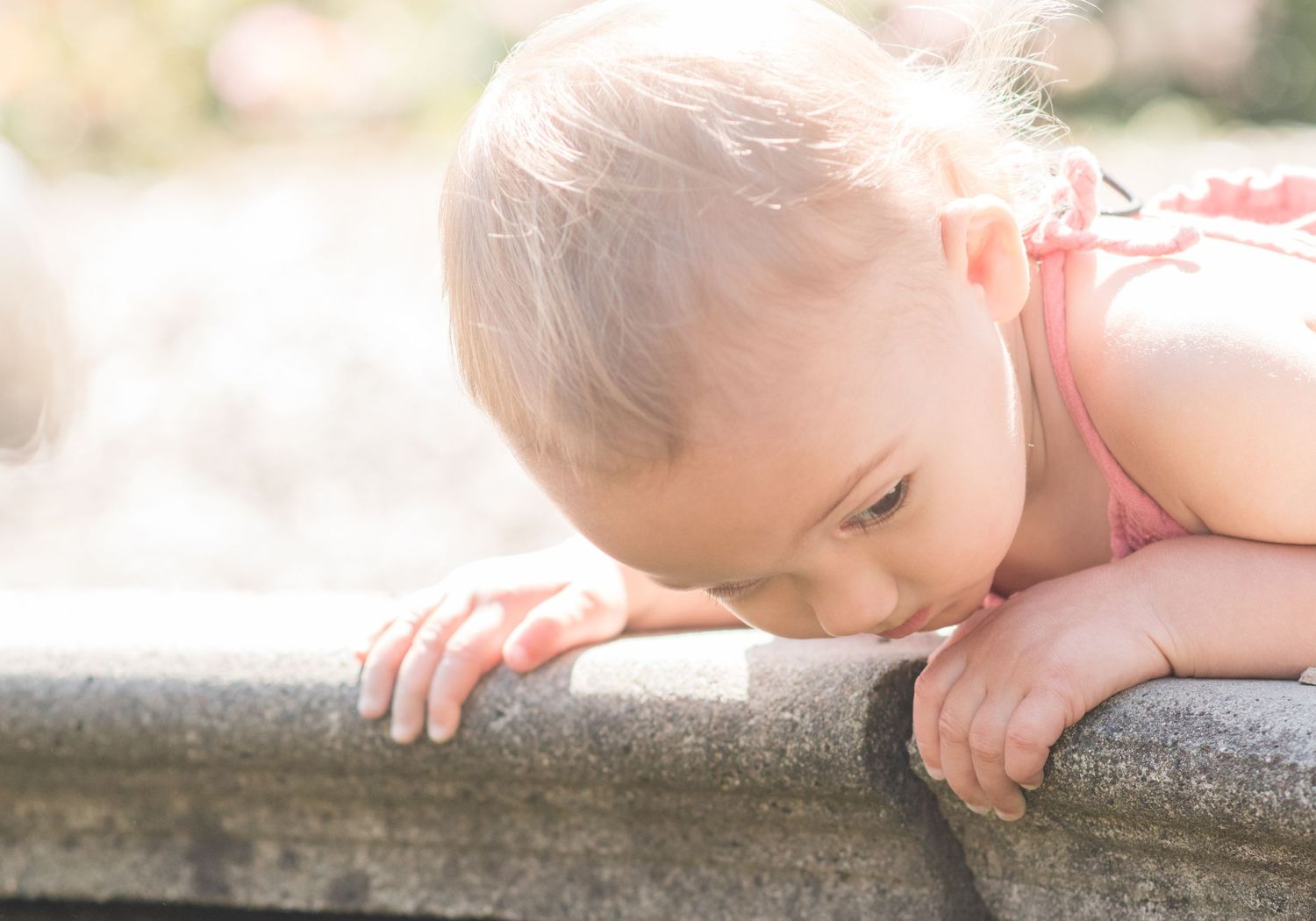 toddler girl looking over into the edge of the fountain. Her hair is reflected in the sunlight. wearing a pink jumper.