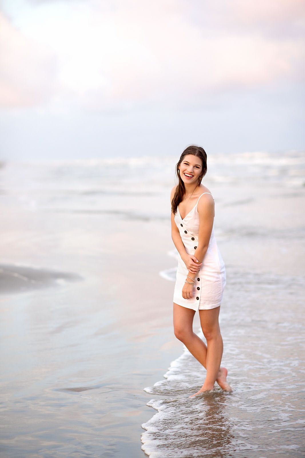 A smiling young woman in a white sundress stands barefoot in ocean waves at sunset.