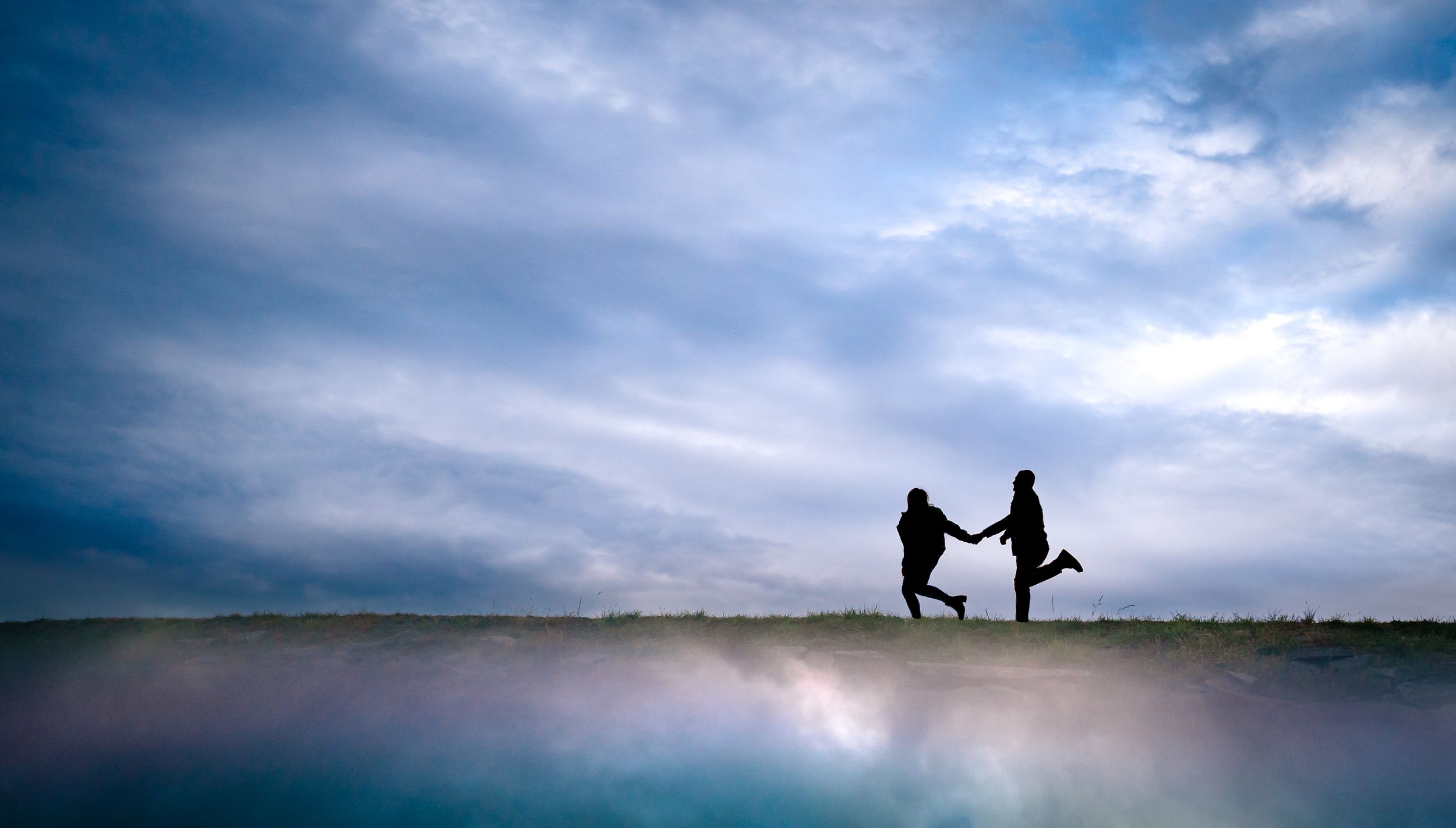 Silhouettes against a dramatic cloudy sky near a lake during sunset.