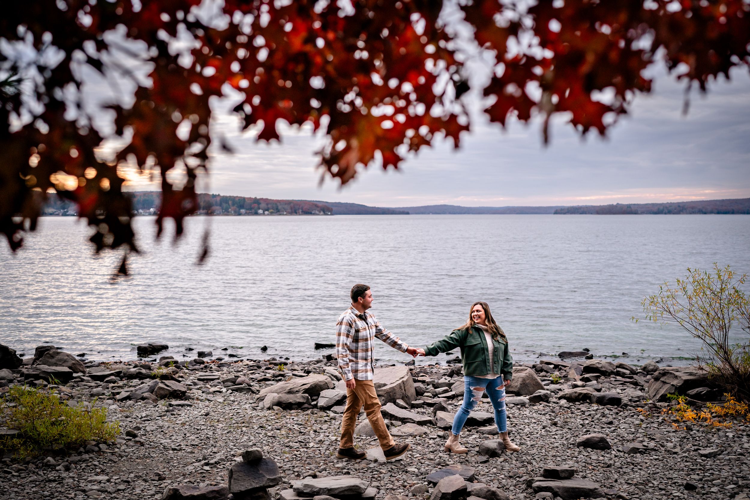 Two people explore a rocky lakeside beach with fall foliage and cloudy skies in the background.