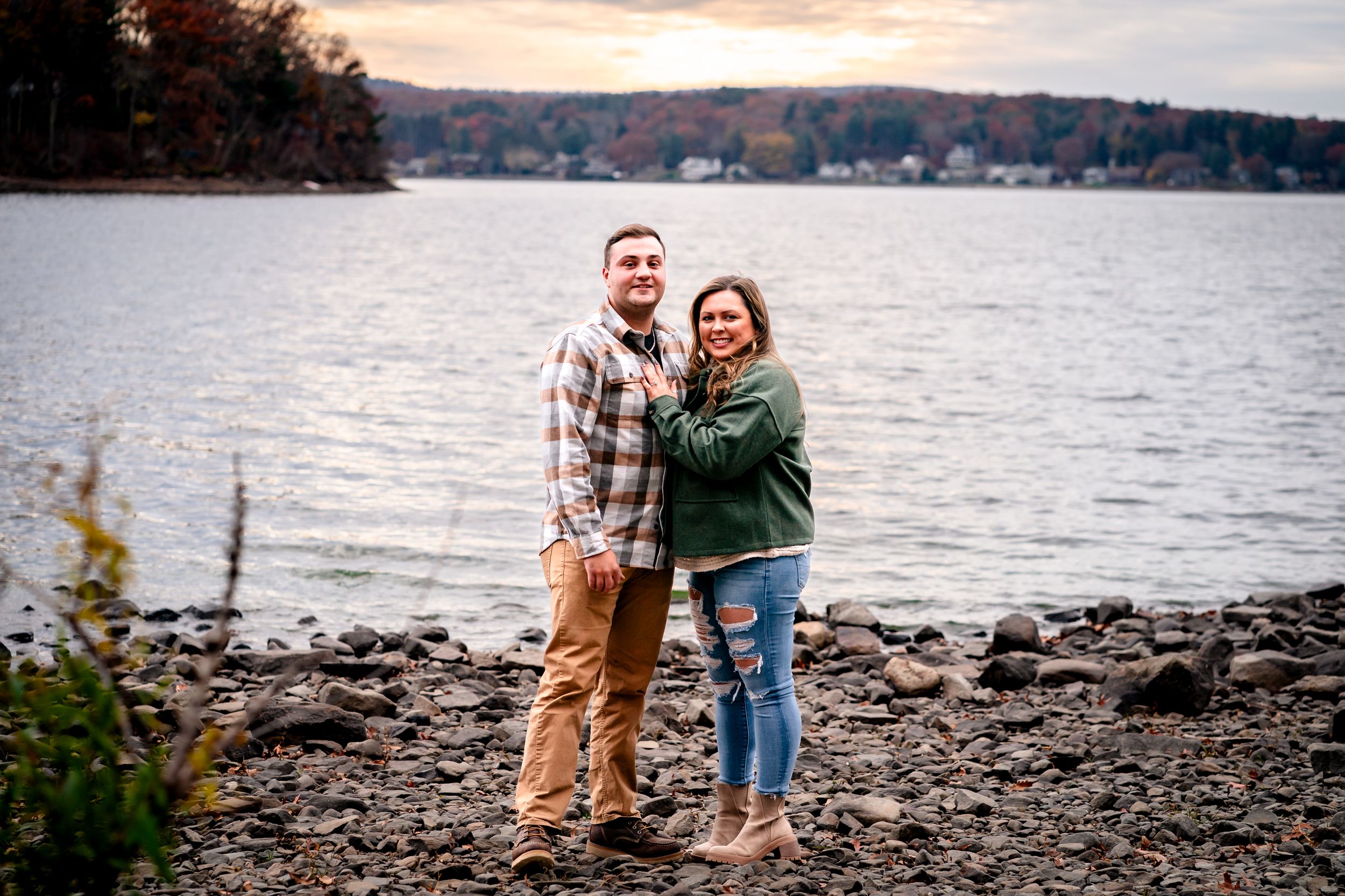 Romantic lakeside engagement photos on rocky beach with sunset backdrop.