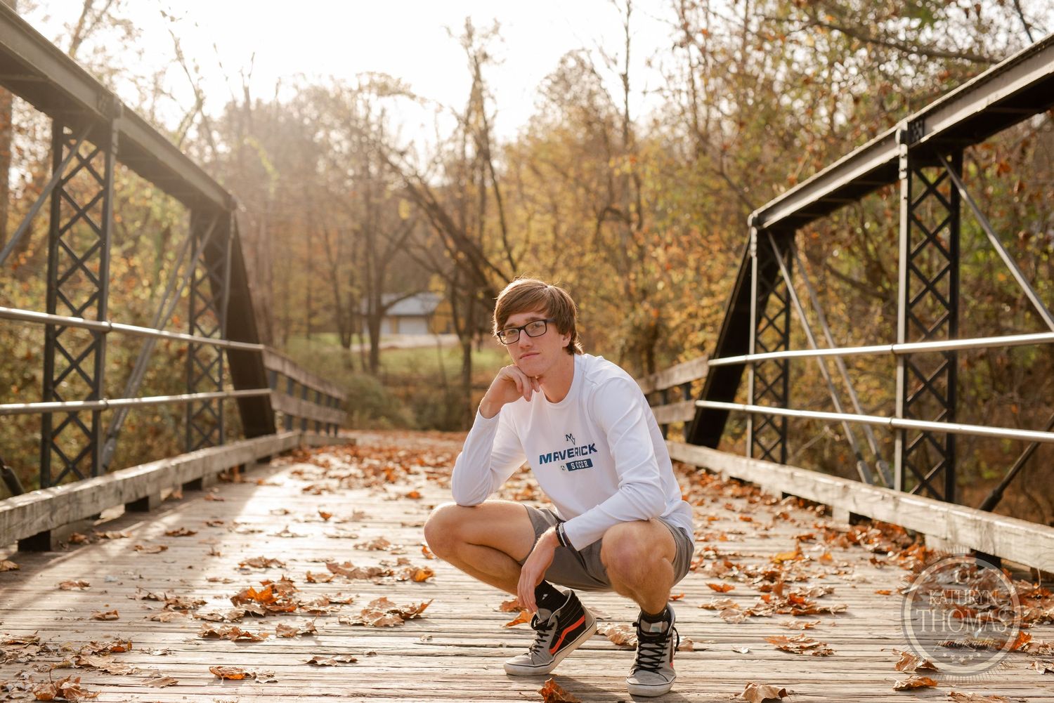 A person crouches on a rustic bridge covered in fallen leaves during autumn.