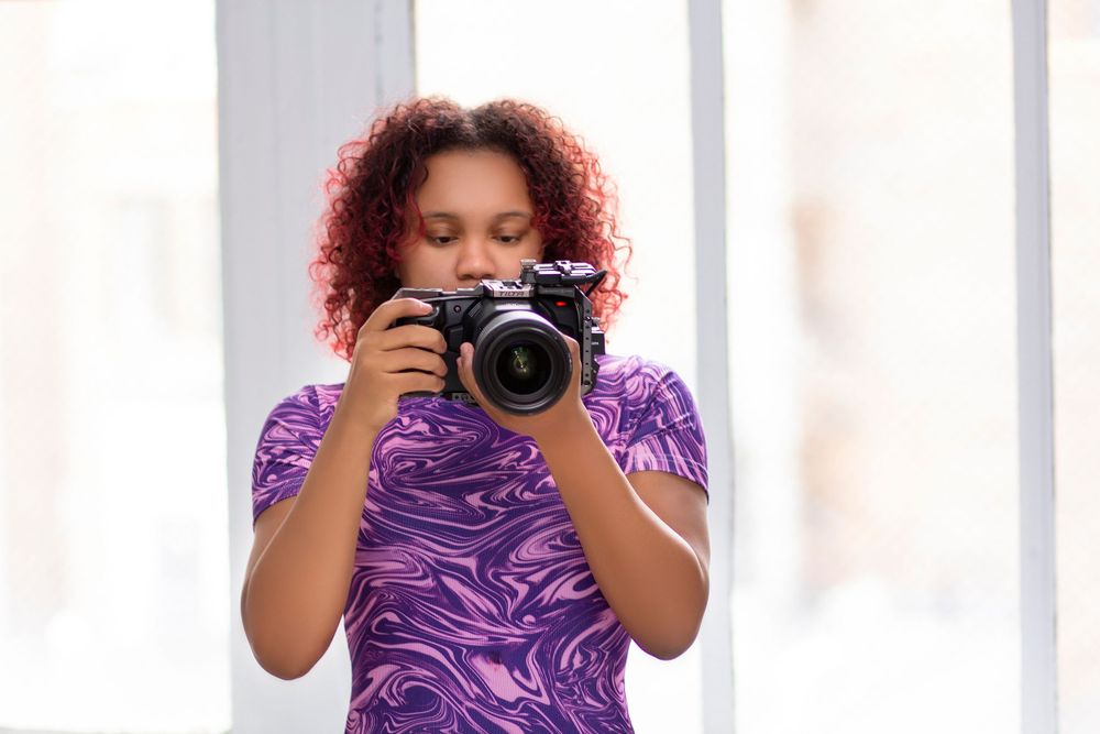 A photographer in a purple dress stands in front of white columns while taking photos with a professional camera.