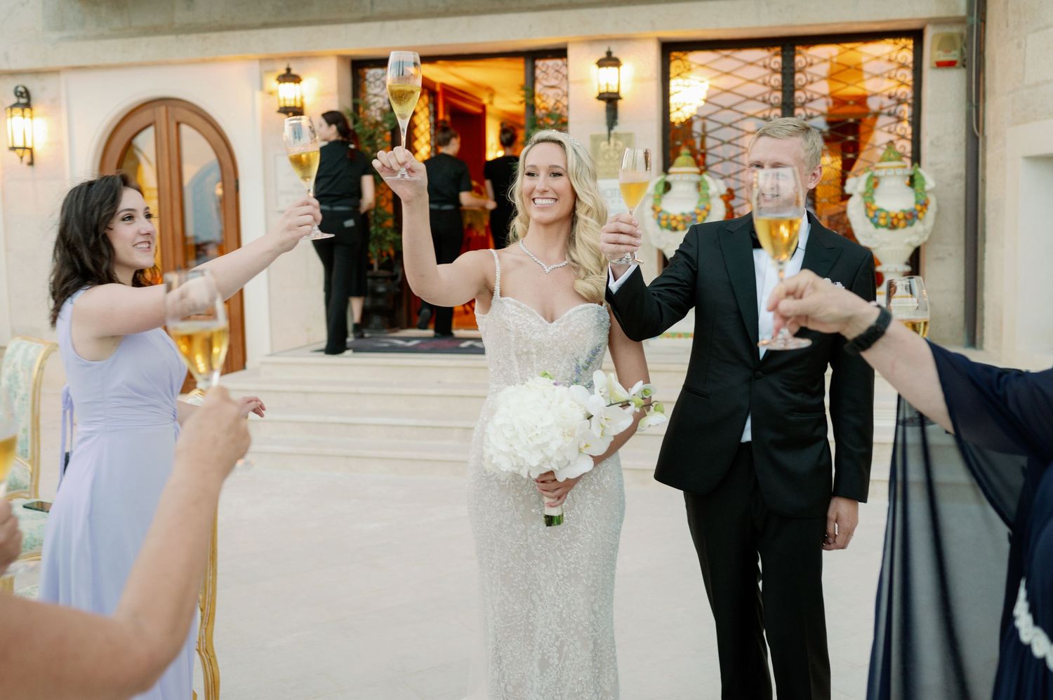 Wedding party celebrating with champagne toast outside elegant venue entrance.