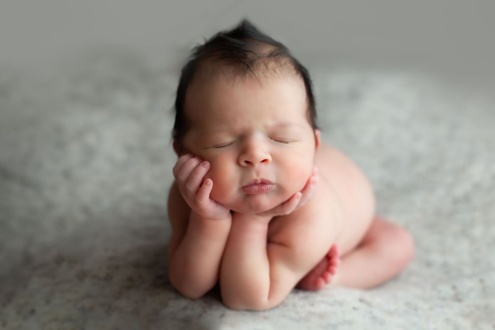Newborn baby sleeping peacefully with hands under chin in classic froggy pose on soft gray blanket.