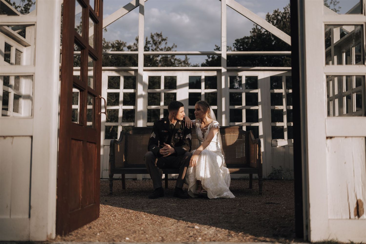 A couple sits together on a bench framed by white wooden doors during an outdoor wedding ceremony.