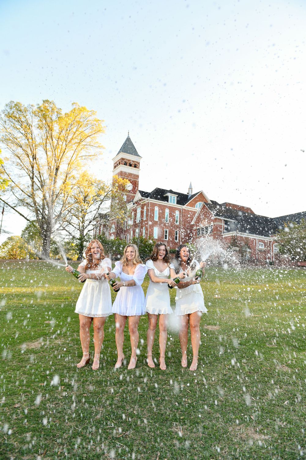 A celebratory moment captured with champagne spraying on a college lawn with a brick clocktower building visible.
