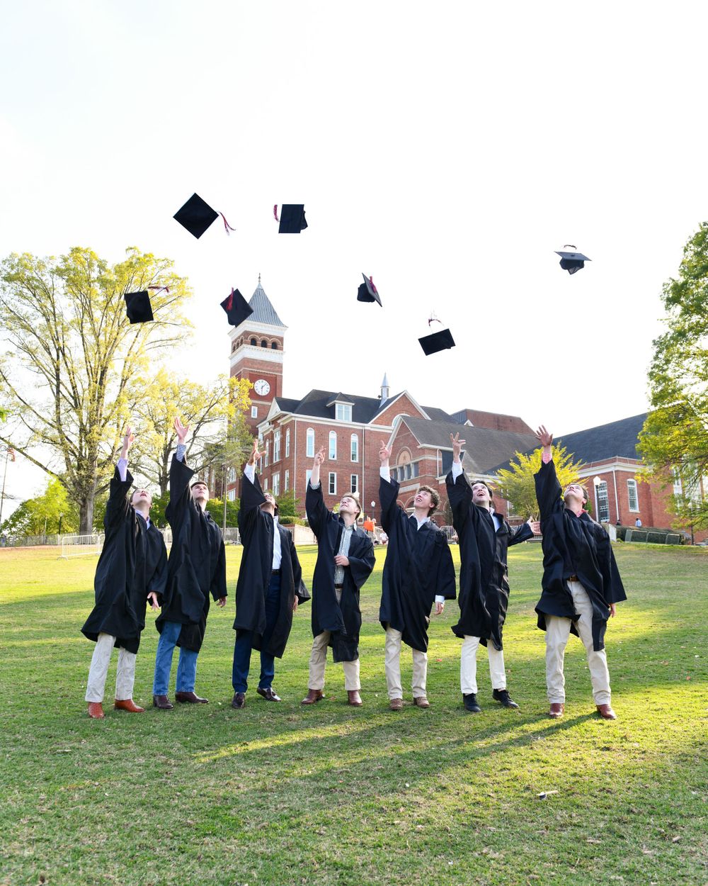 Graduates in black robes throw their caps in the air in celebration on a lawn with a brick building behind them.