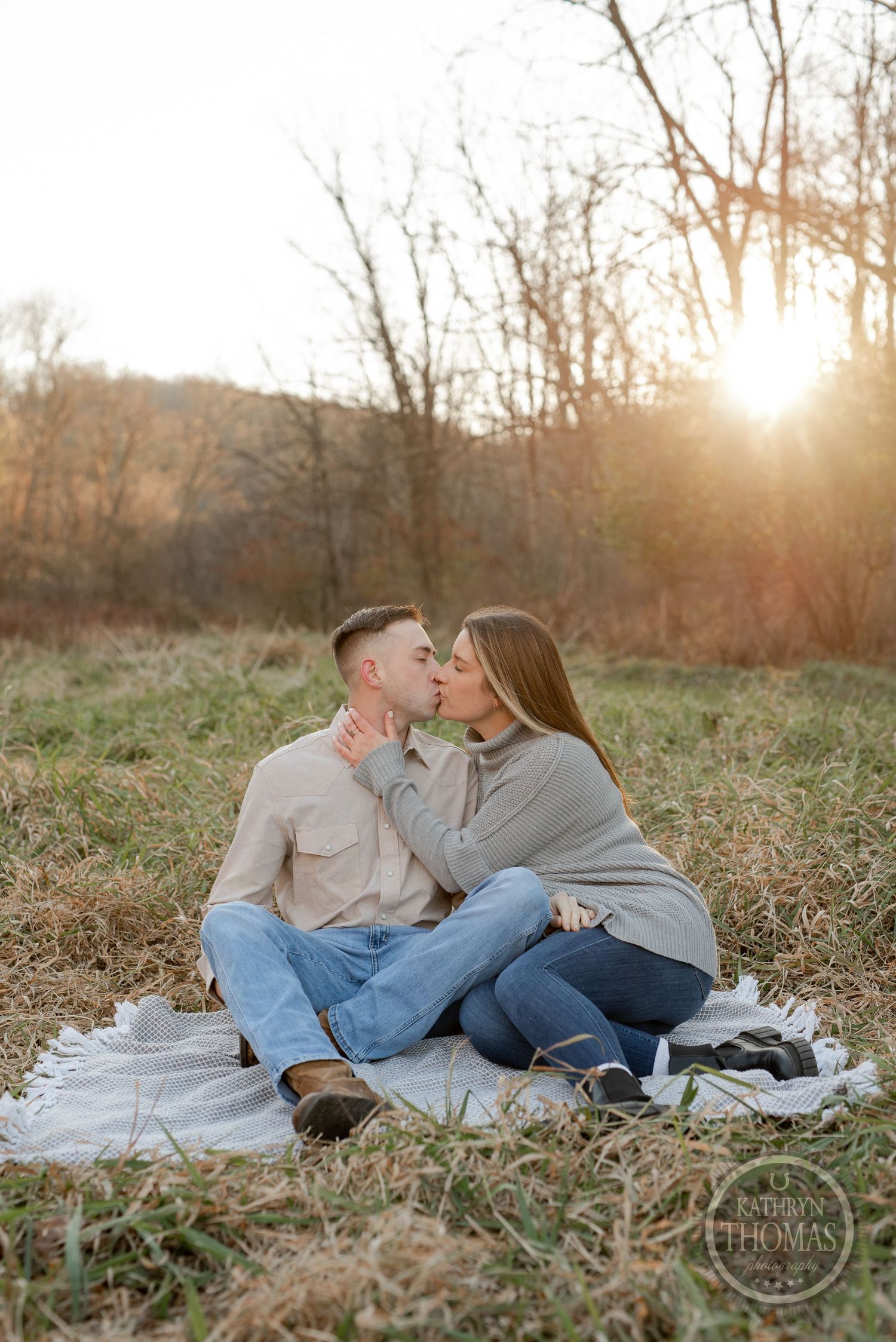 A couple shares a tender moment while sitting on a white blanket in a sunlit autumn field.