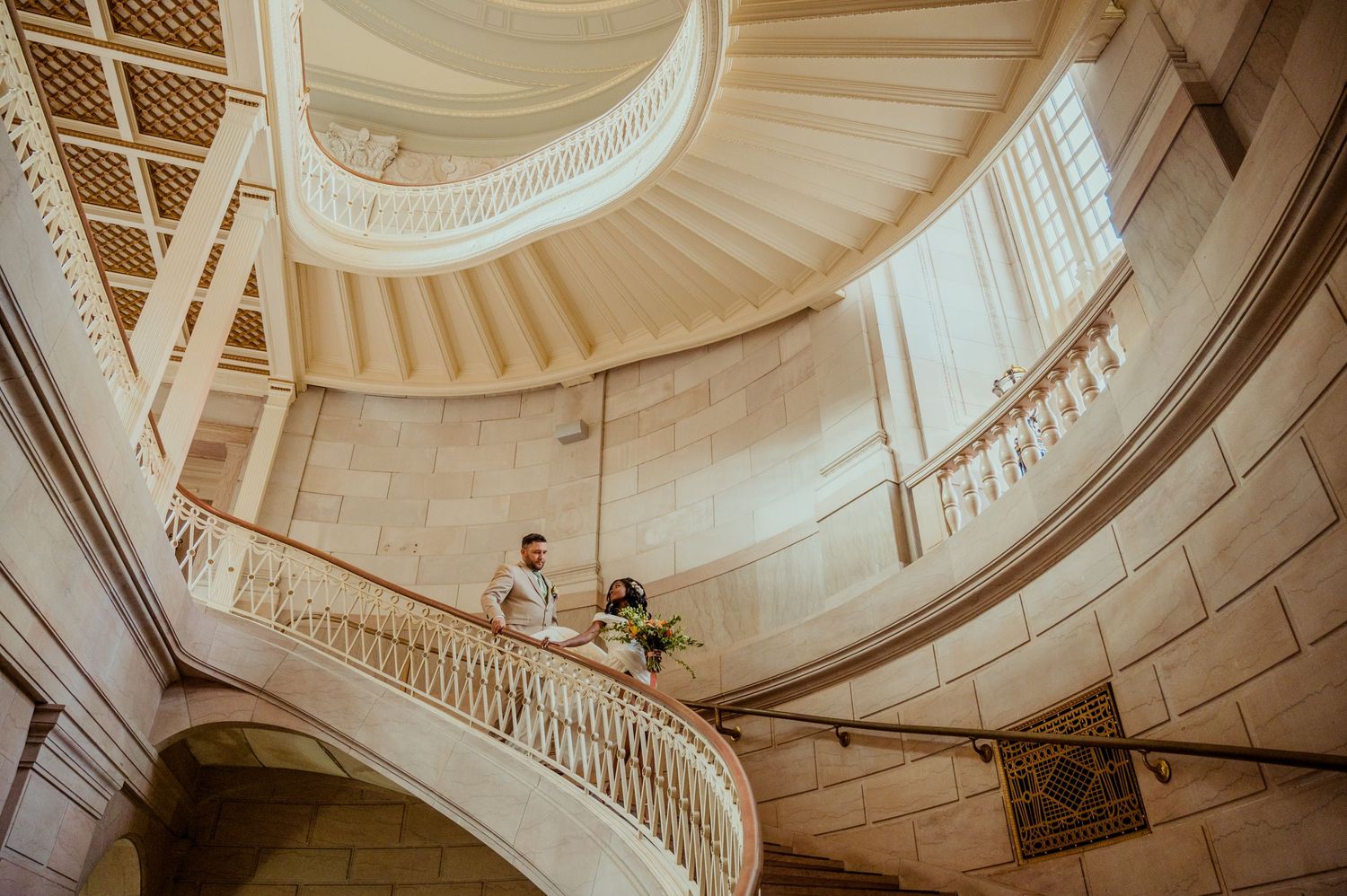 A couple poses on an elegant curved white marble staircase in a grand building.