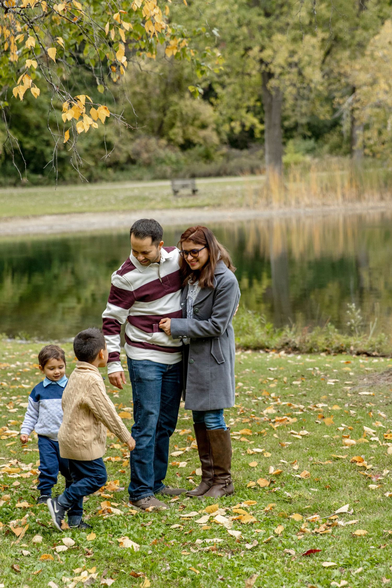 Family enjoying an autumn day by a pond with fallen yellow leaves on grass and reflective water in background at Spencer park in Rochester