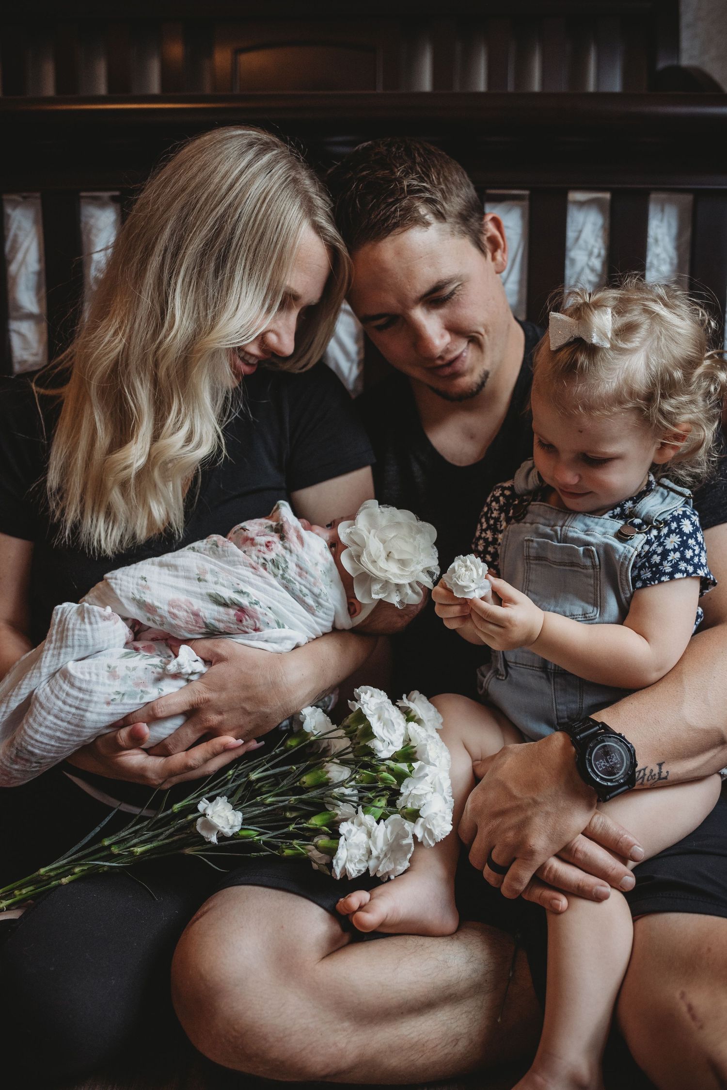 A family sits together on a dark couch with white flowers, sharing an intimate moment with their newborn and toddler.