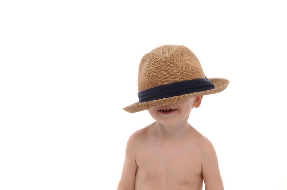 Child wearing brown fedora hat smiling playfully against white backdrop.