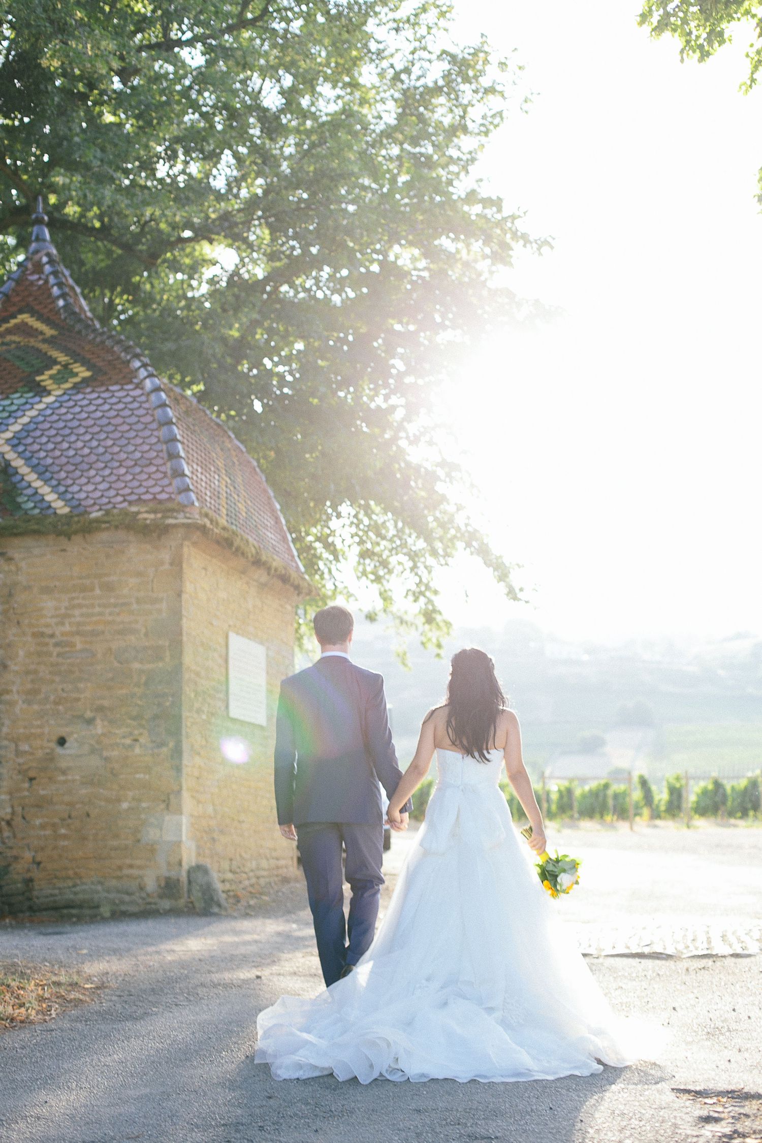 Newlyweds walking hand in hand near rustic building during golden hour.