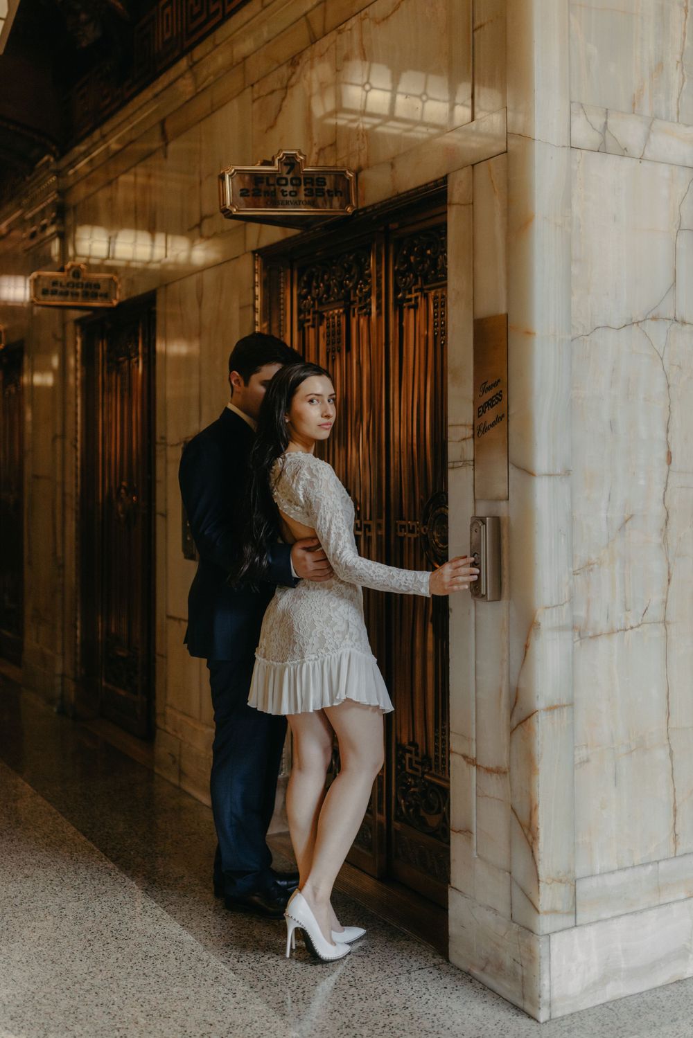 Couple in formal wear pose intimately by vintage elevator doors in marble hallway.