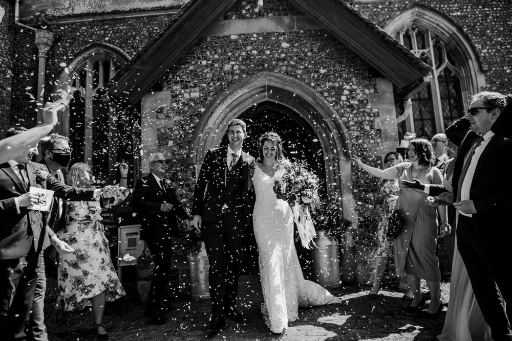 Wedding couple exits church under shower of confetti in black and white photo.