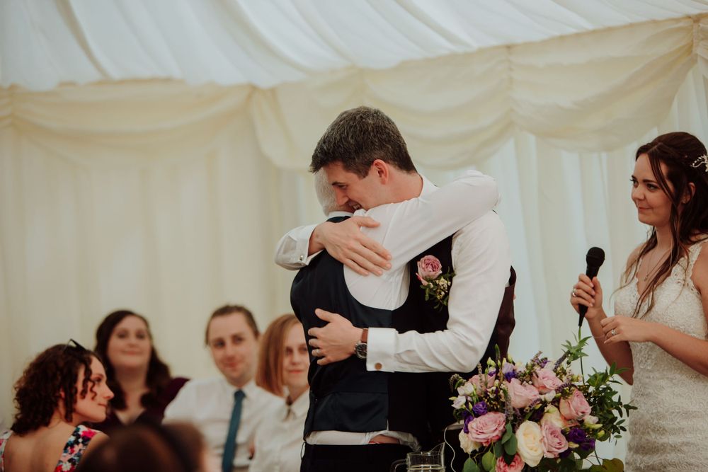 Emotional embrace during wedding reception speech in white marquee tent.