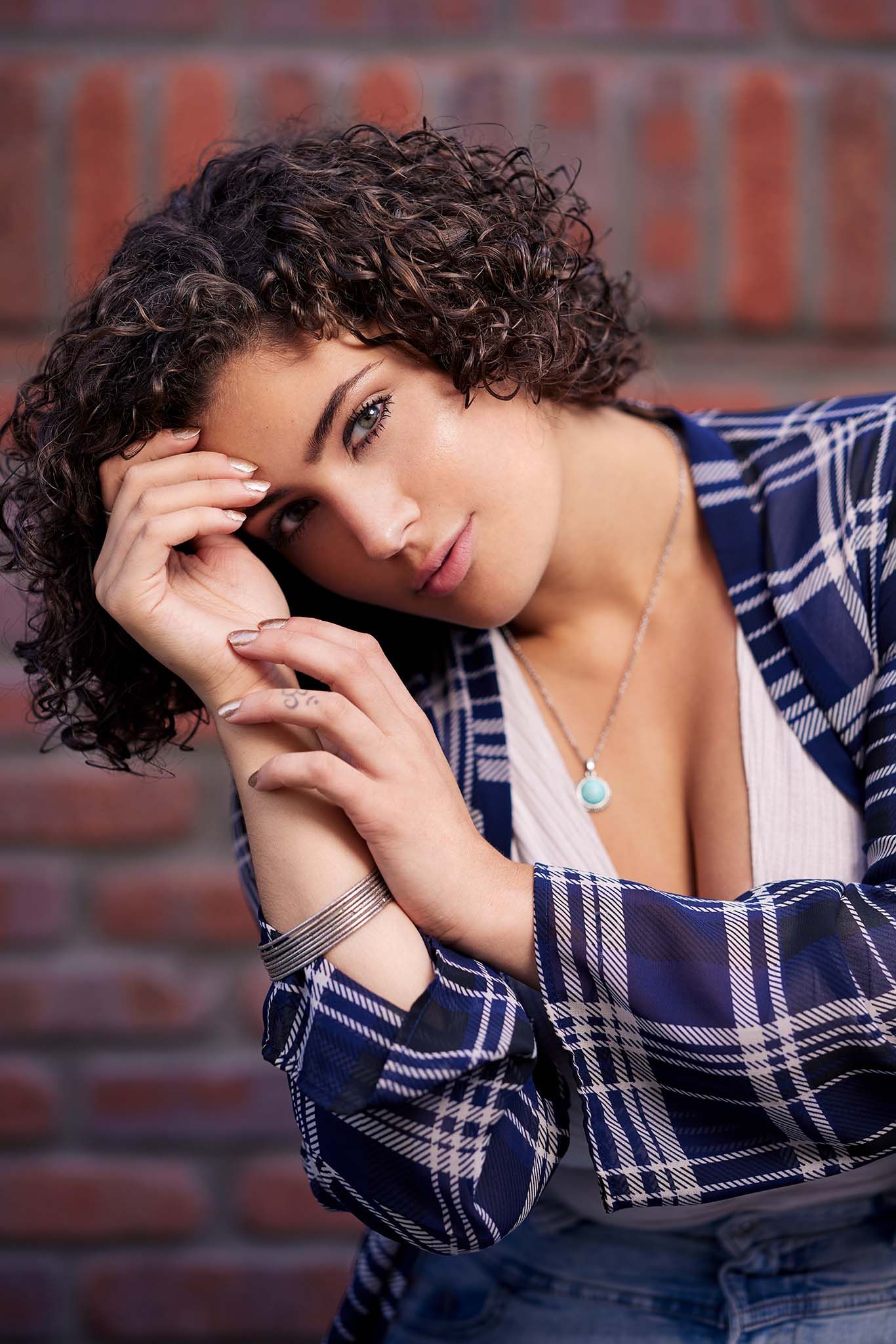 A portrait of a person with curly hair wearing a plaid shirt against a brick wall.
