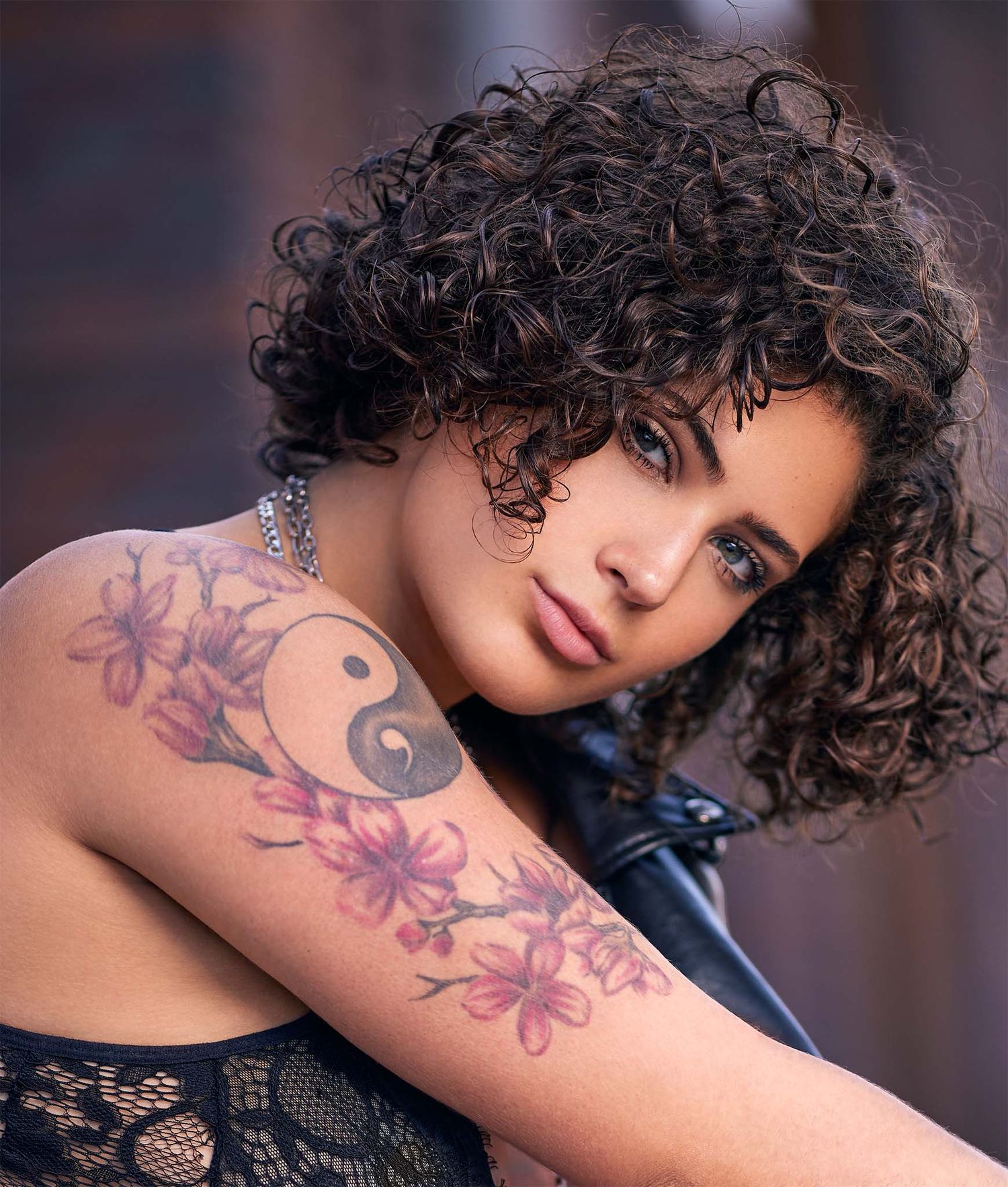 A artistic portrait showcasing floral tattoos and curly hair against a brick background.