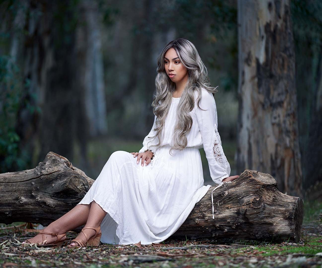 Woman in flowing white dress sitting on fallen log in enchanted forest setting.