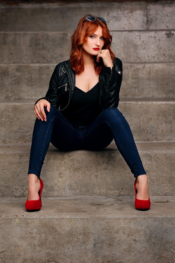 Model in black leather jacket and red heels poses on concrete steps.
