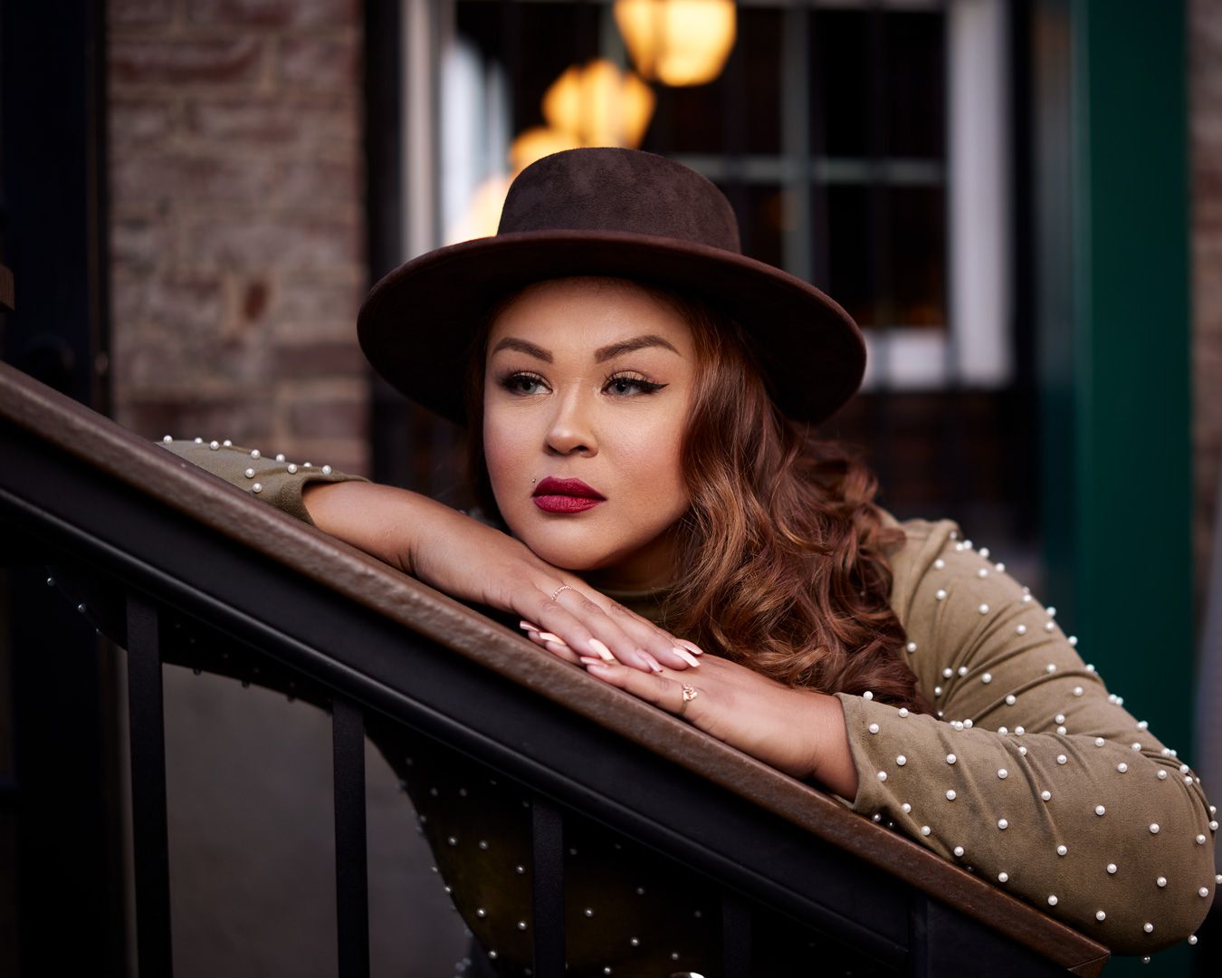 Glamour shot of model in brown hat and pearl-studded dress leaning on railing.