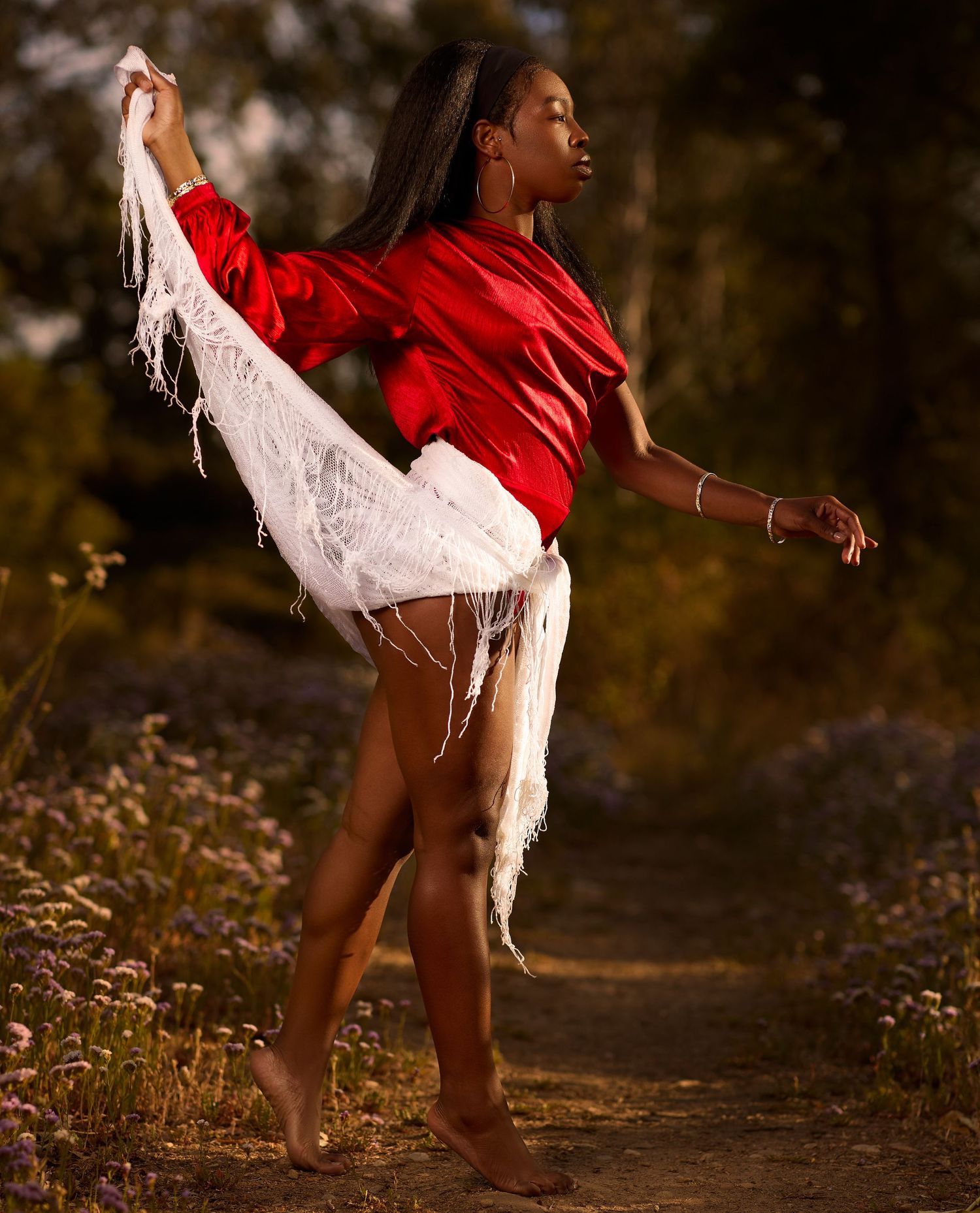 Dancer in red top with flowing white fabric performs on nature trail at golden hour.