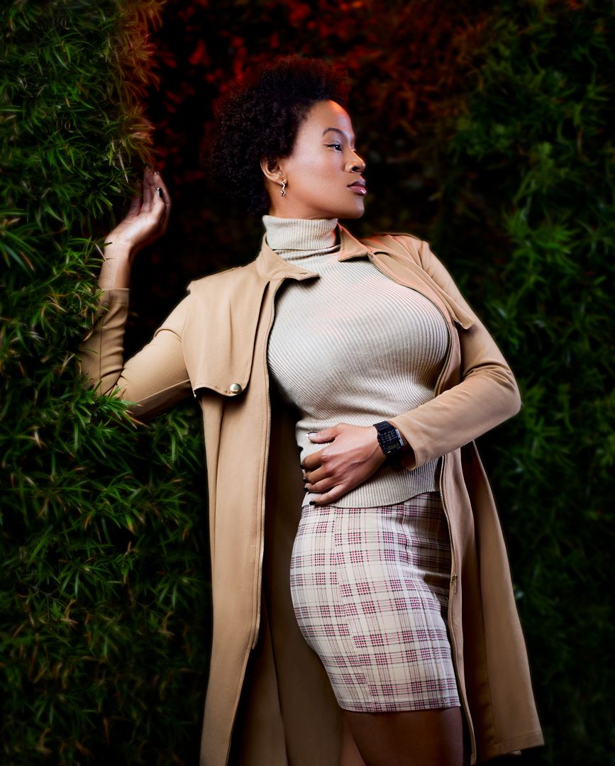 Fashion model in beige coat and plaid skirt poses against green foliage background.