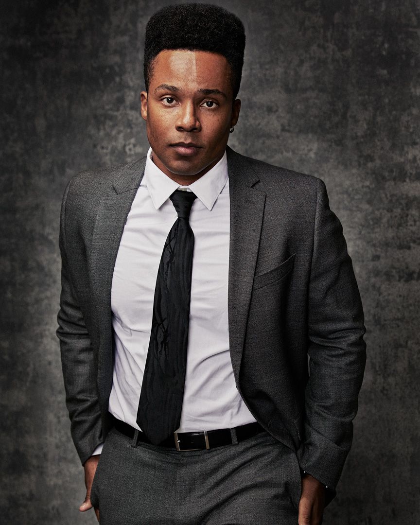 Professional headshot in gray suit and black tie against textured background.