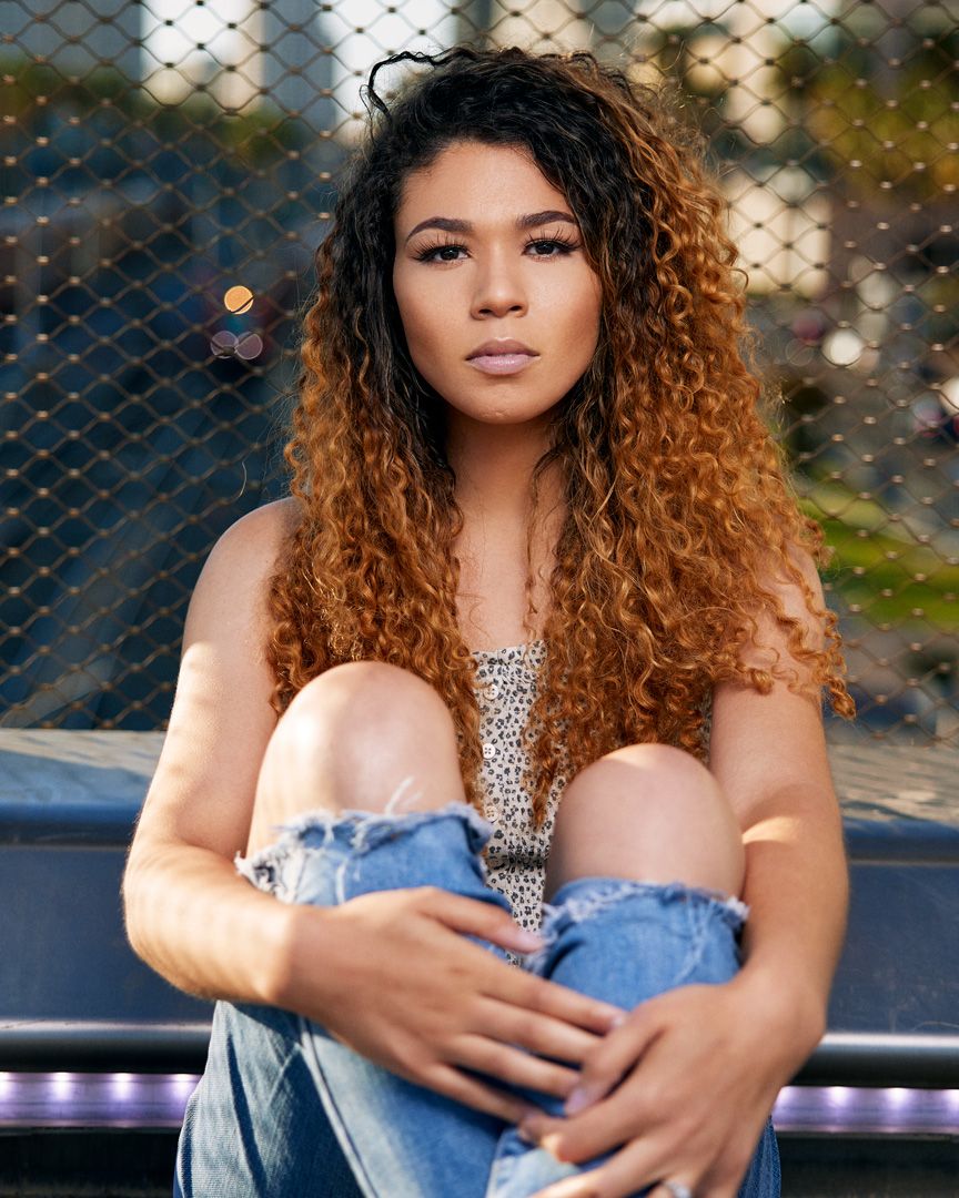 Model with ombre curly hair in sparkly top and denim sits casually outdoors.