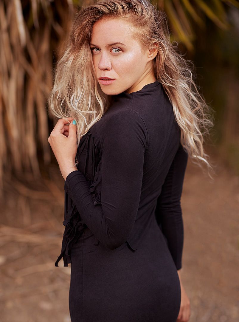 Portrait of woman with windswept blonde hair wearing black dress against natural backdrop.