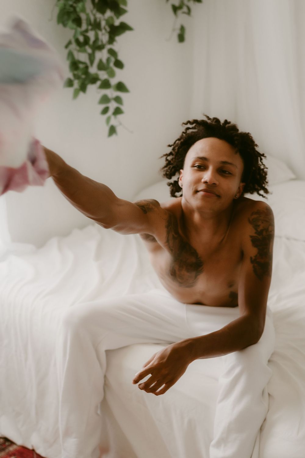 Person lounging on white bedding with hanging plants in a bright, minimalist bedroom setting.