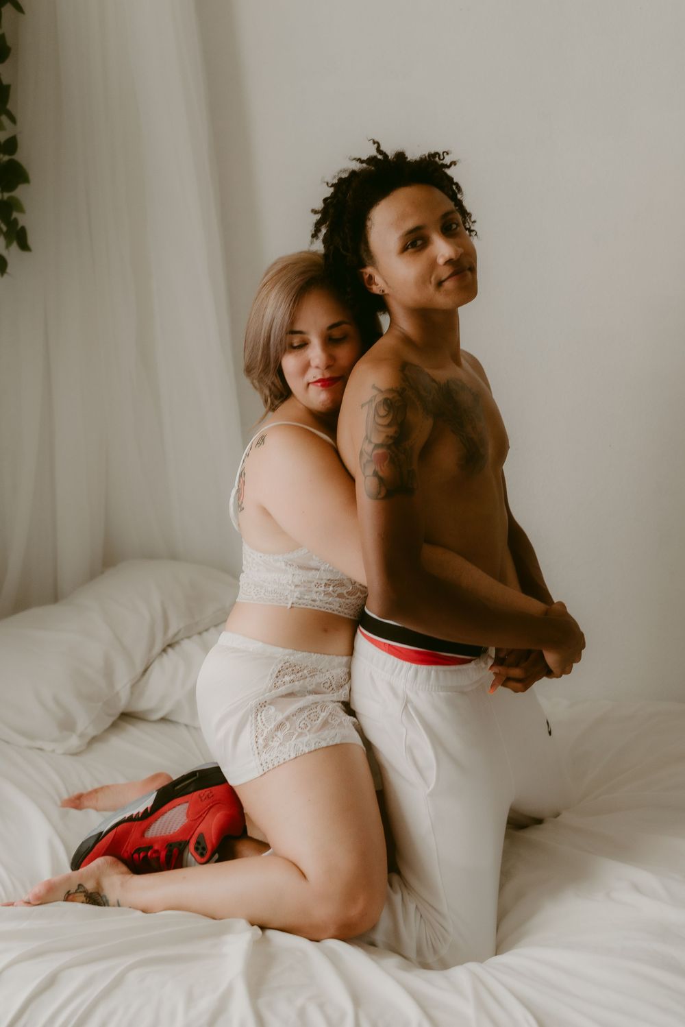 Two people in casual intimate poses on white bedding during a cozy photoshoot.