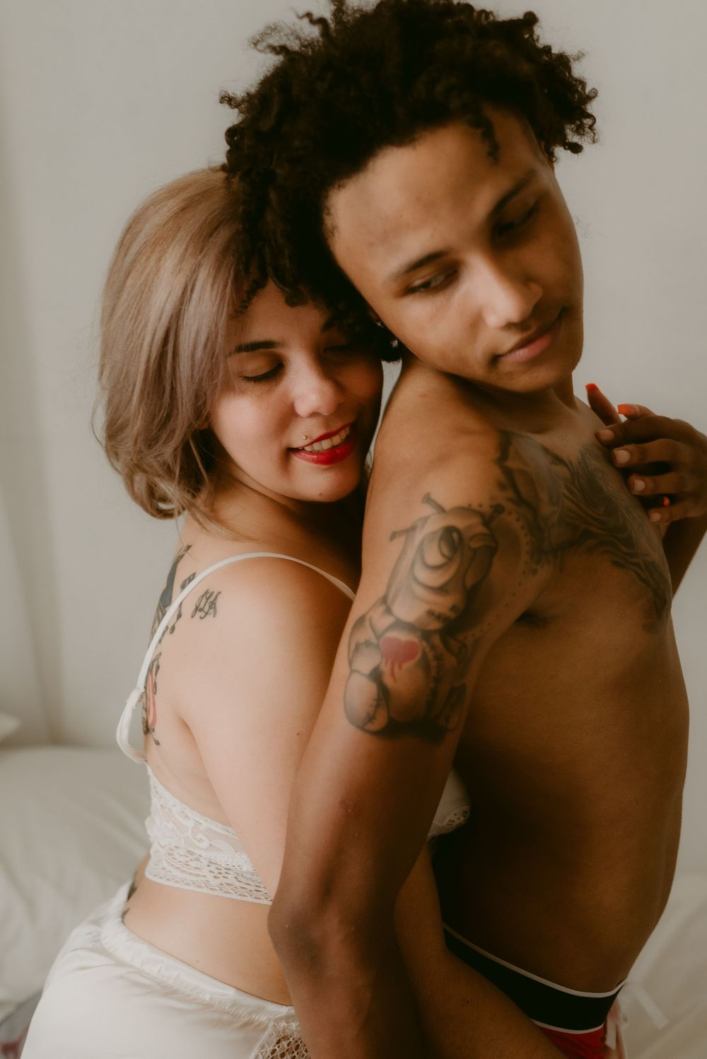 Intimate couple in white lingerie embracing while showing tattoos in a soft natural light setting.