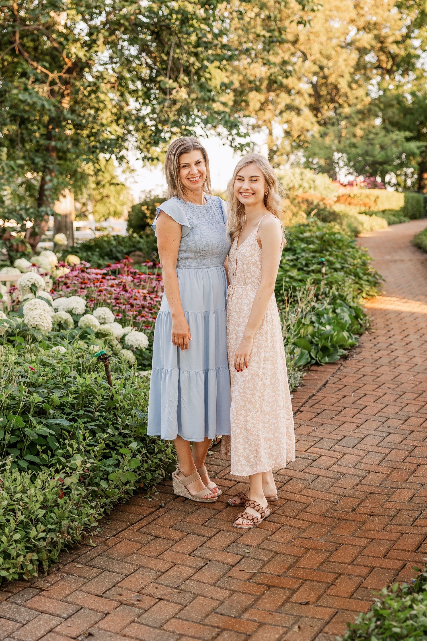 Two people in summer dresses pose on a brick path surrounded by blooming garden flowers.