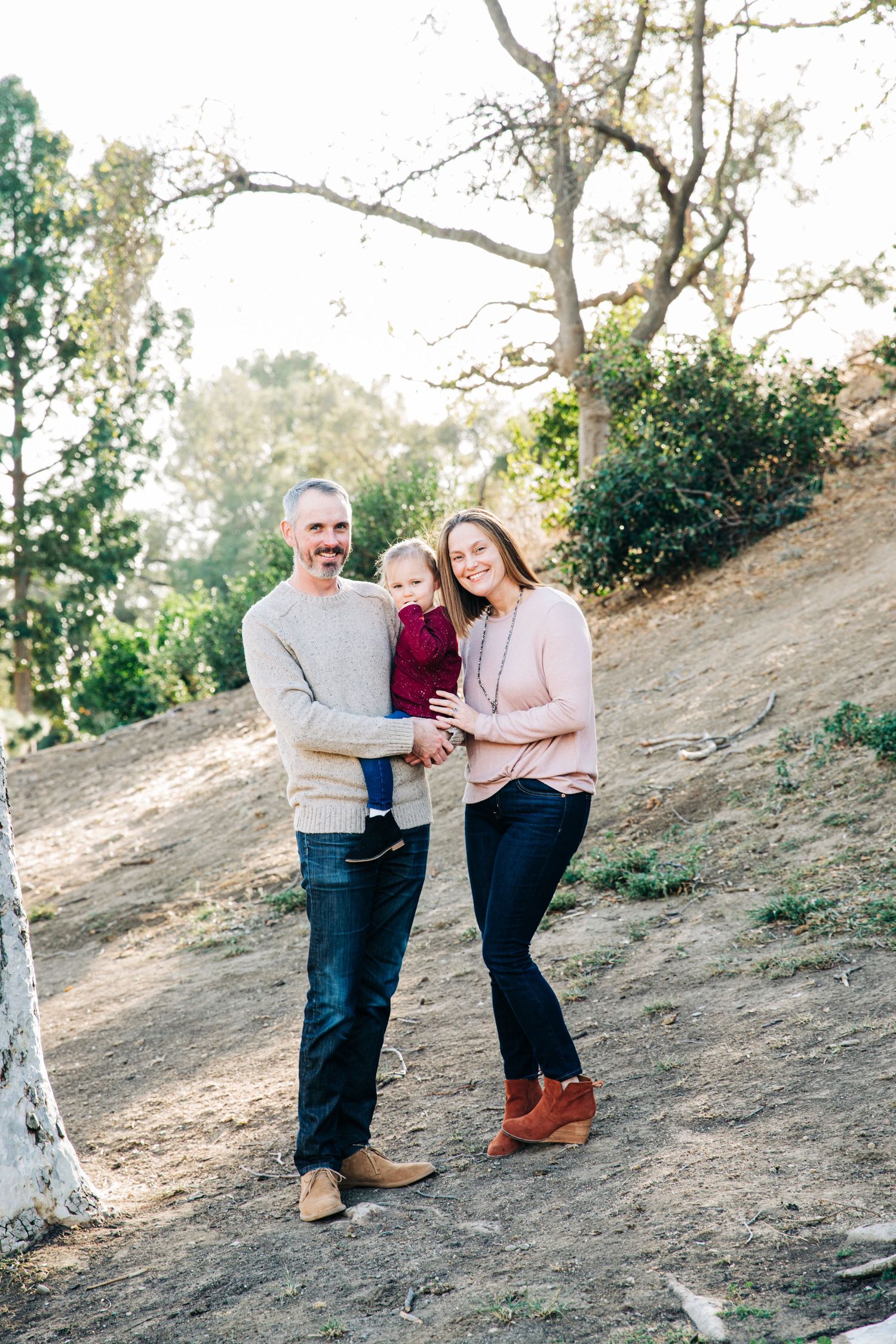 A couple shares a tender moment while standing on a hillside path surrounded by trees and natural lighting.