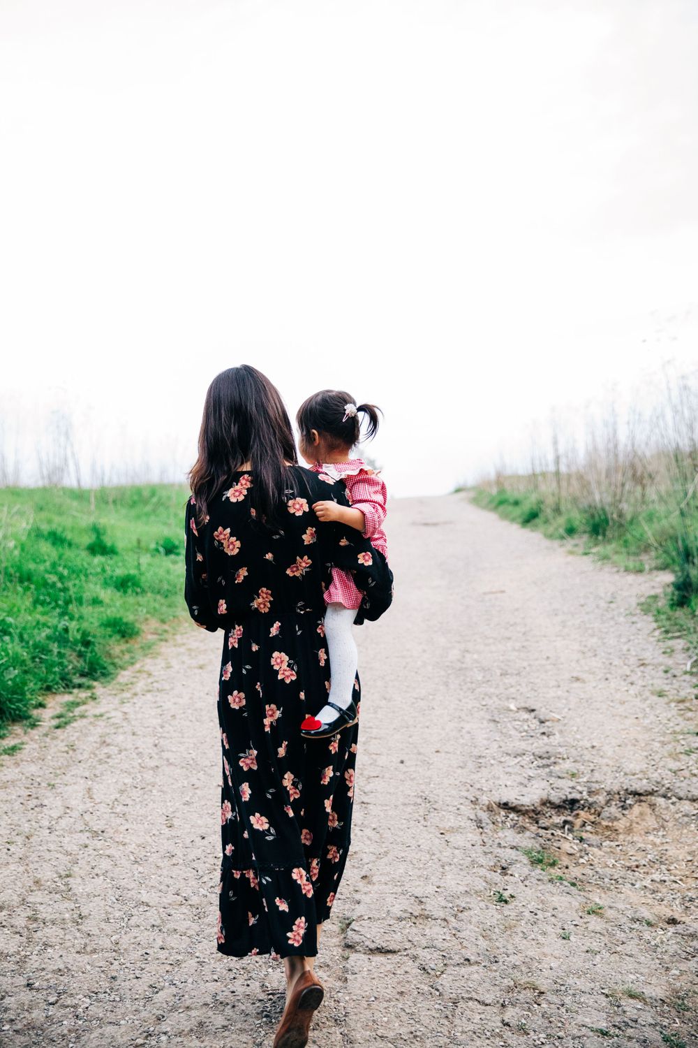 A woman in a floral dress carries a small child while walking down a dirt path.