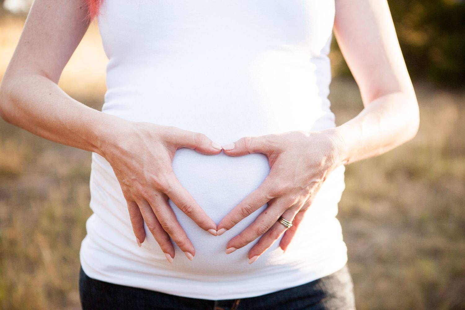 Hands forming a heart shape over a pregnant belly in white shirt against sunset background.