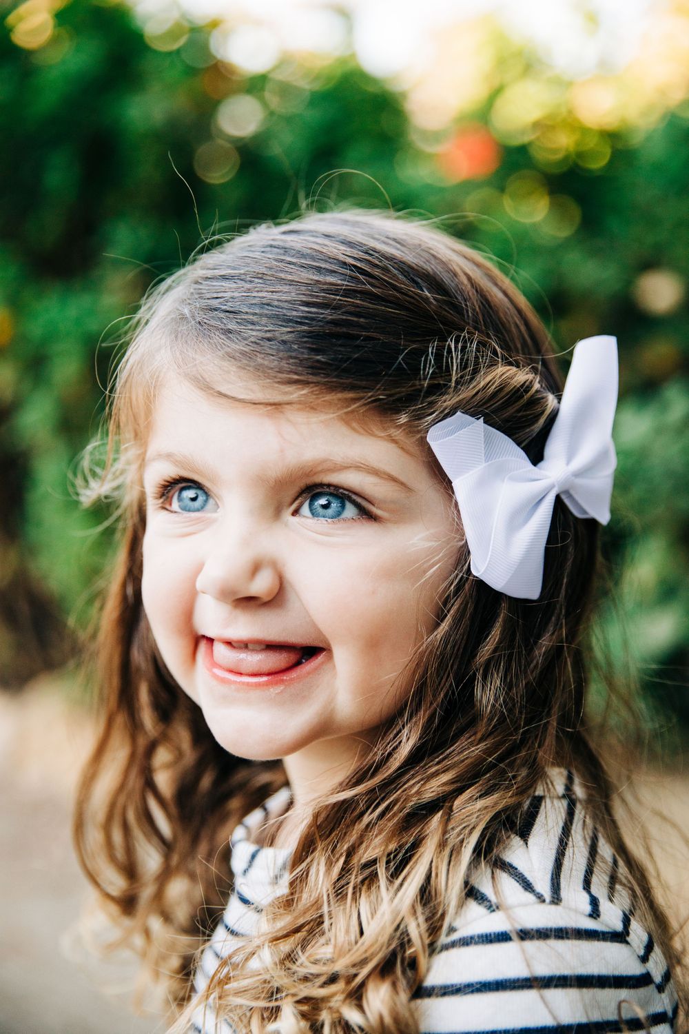 A child wearing a white bow and striped shirt smiles against a blurred green background.